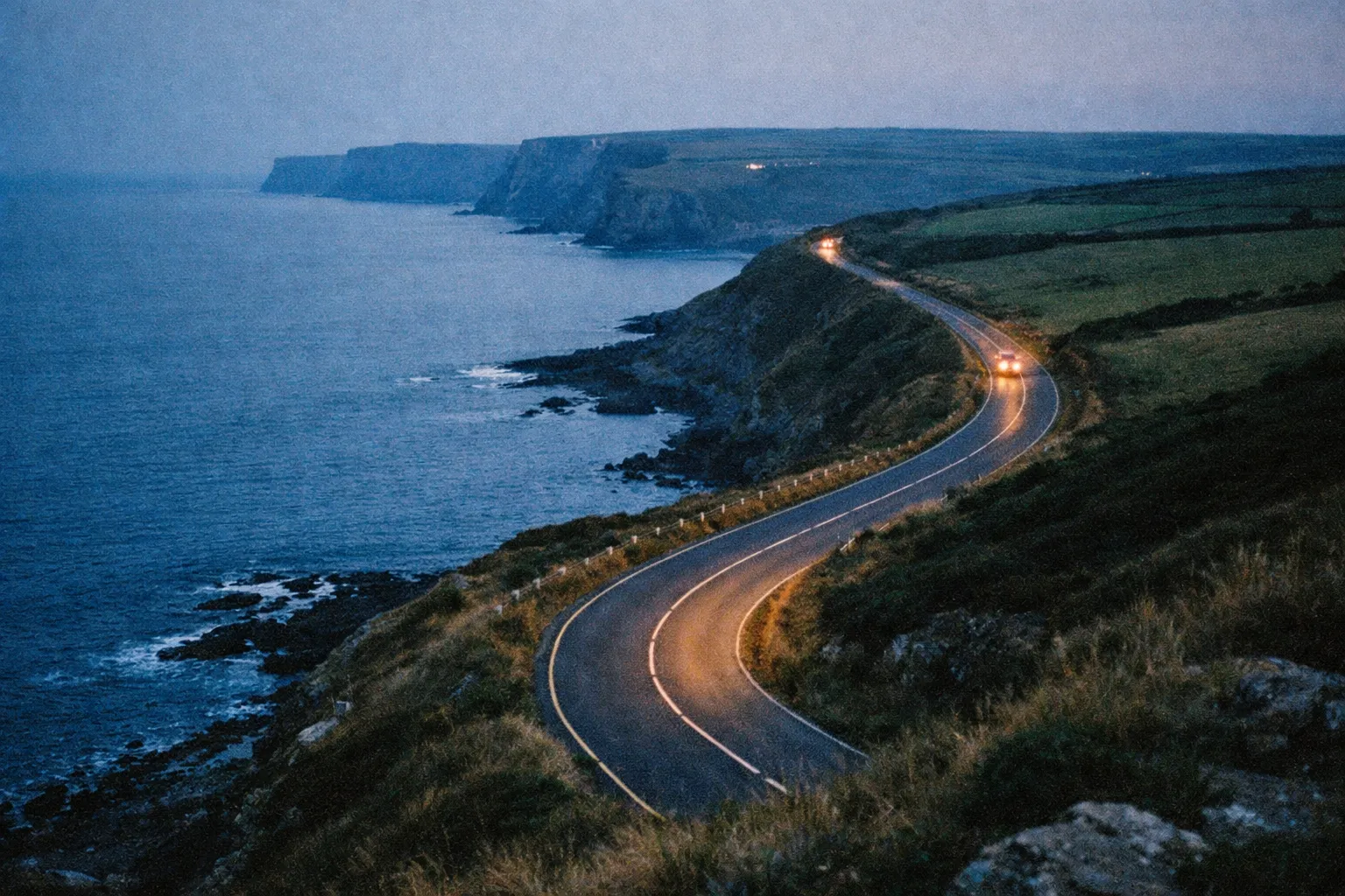 A winding coastal road with cars overlooks dramatic cliffs and the Atlantic Ocean at dusk.