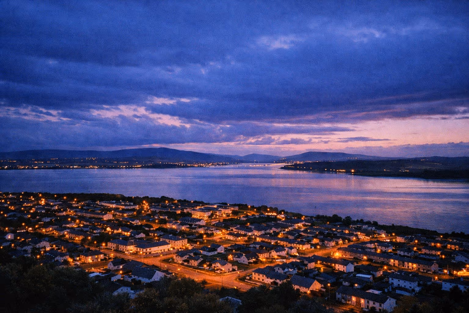 A riverside town along the Shannon estuary at dusk with lights glowing and hills in the background.