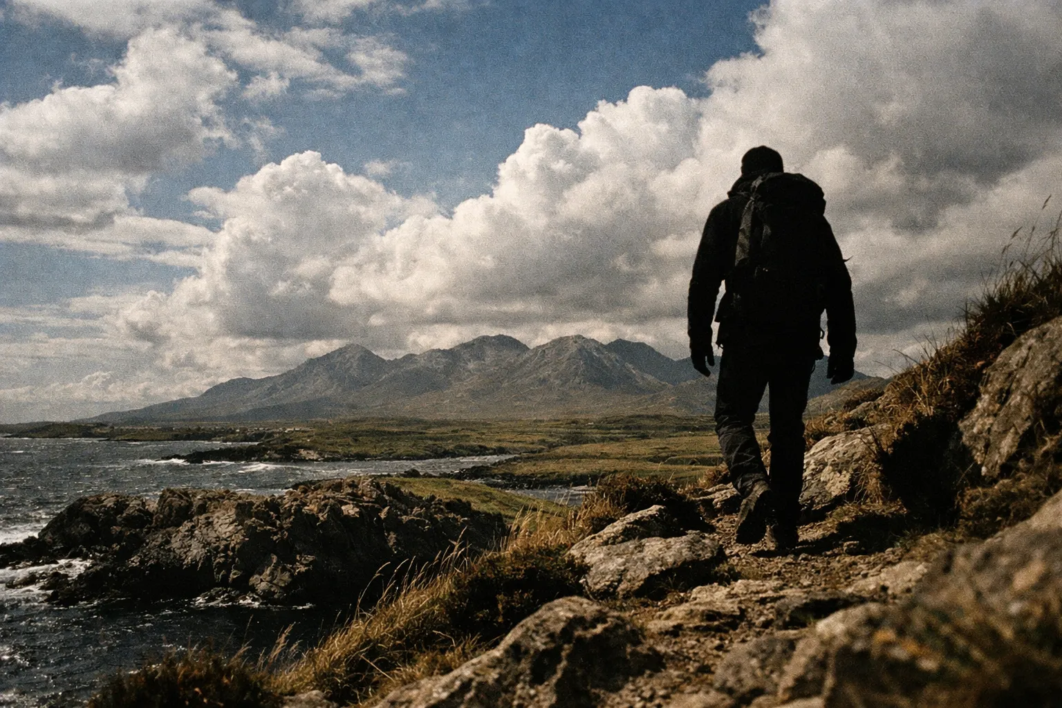 A hiker walks along a rocky coastal path with mountains in the background in Connemara, Ireland.
