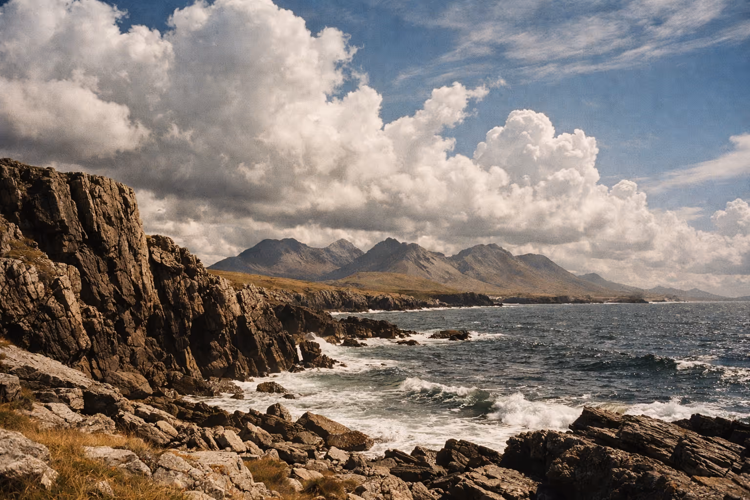 Rocky Atlantic coastline with grassy cliffs and mountains in the distance under a partly cloudy sky in Connemara, Ireland.