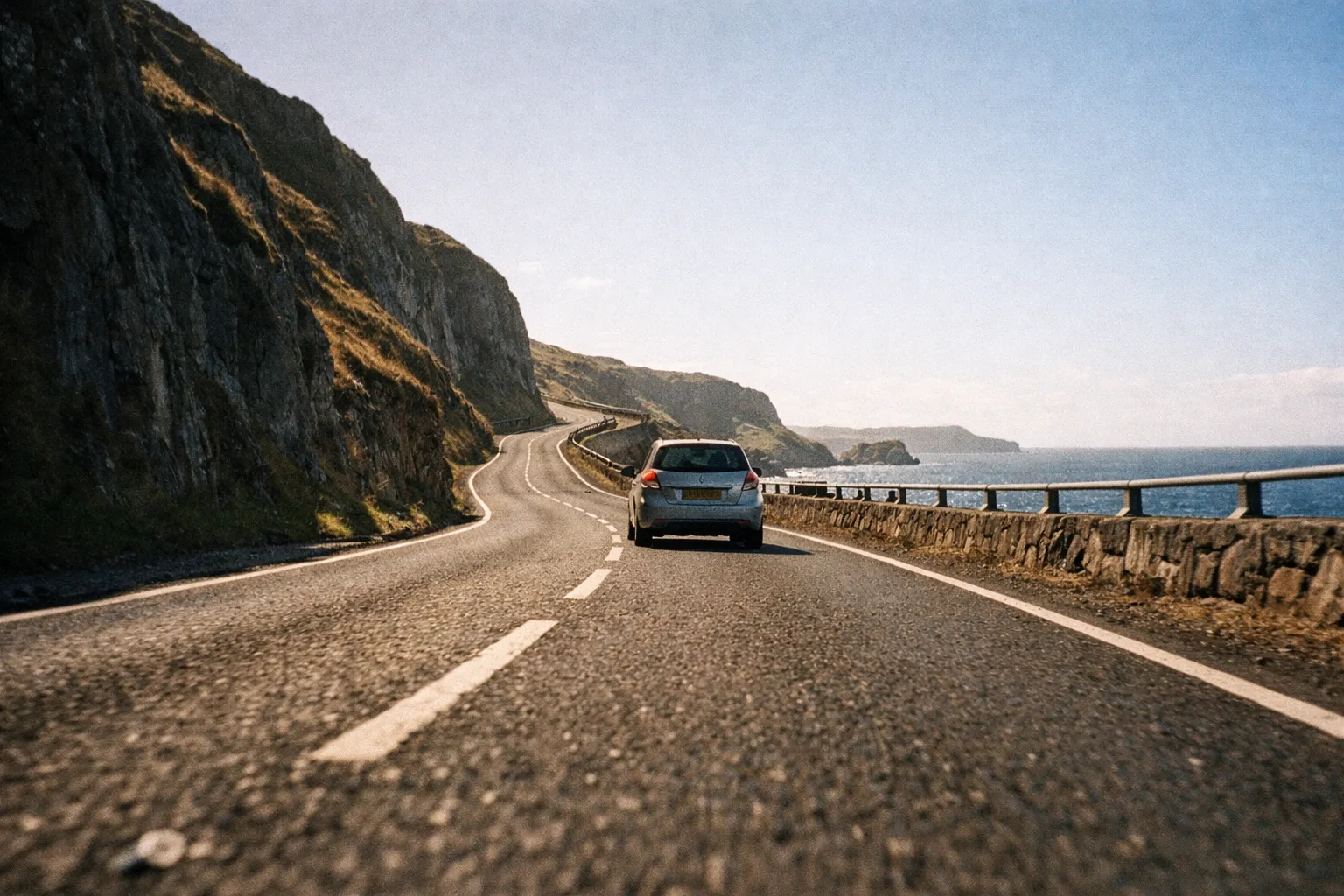 A car drives along a coastal road beside cliffs with the sea visible to the right.