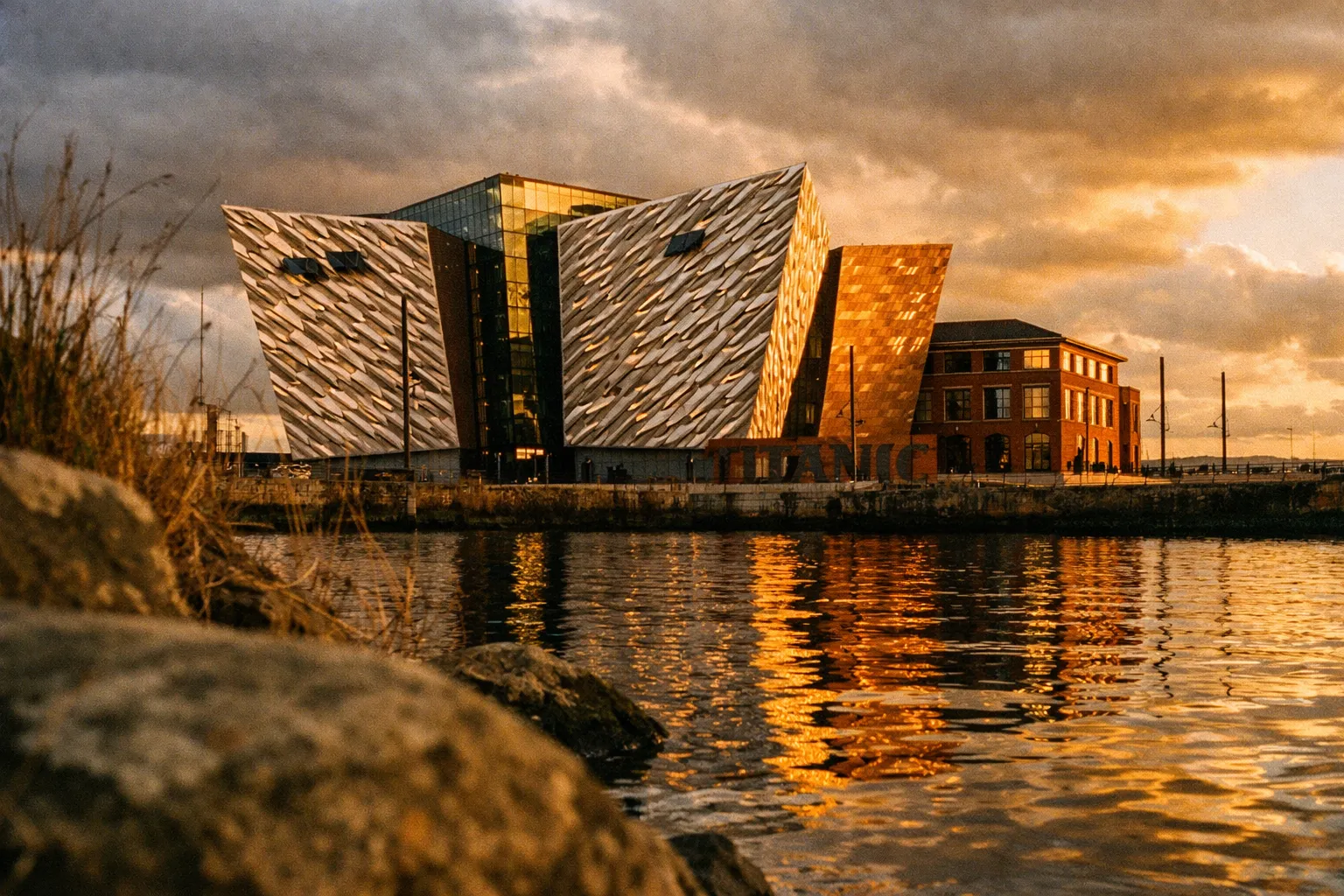 The Titanic Belfast museum stands beside a reflective waterfront under a cloudy evening sky.