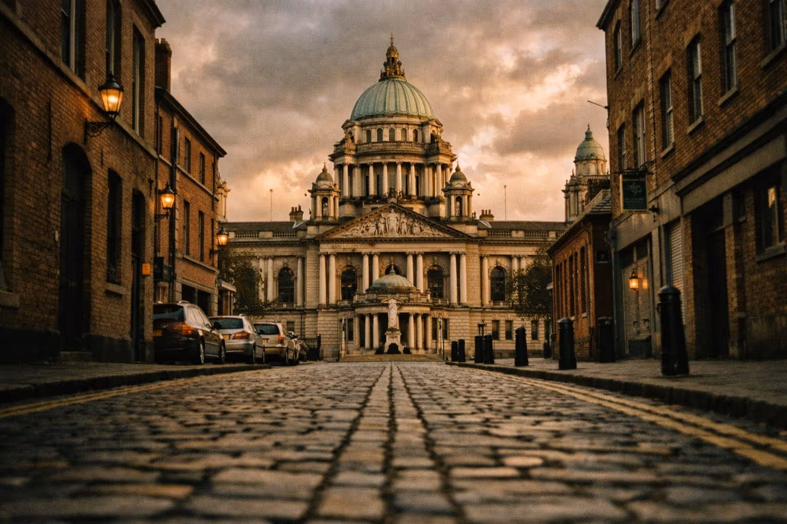 Belfast City Hall viewed from a cobbled street lined with brick buildings at dusk.