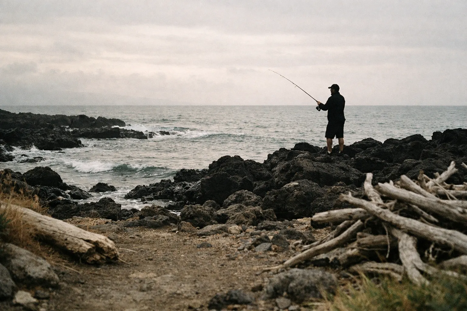 A person fishing from black volcanic rocks by the ocean shore under cloudy skies.