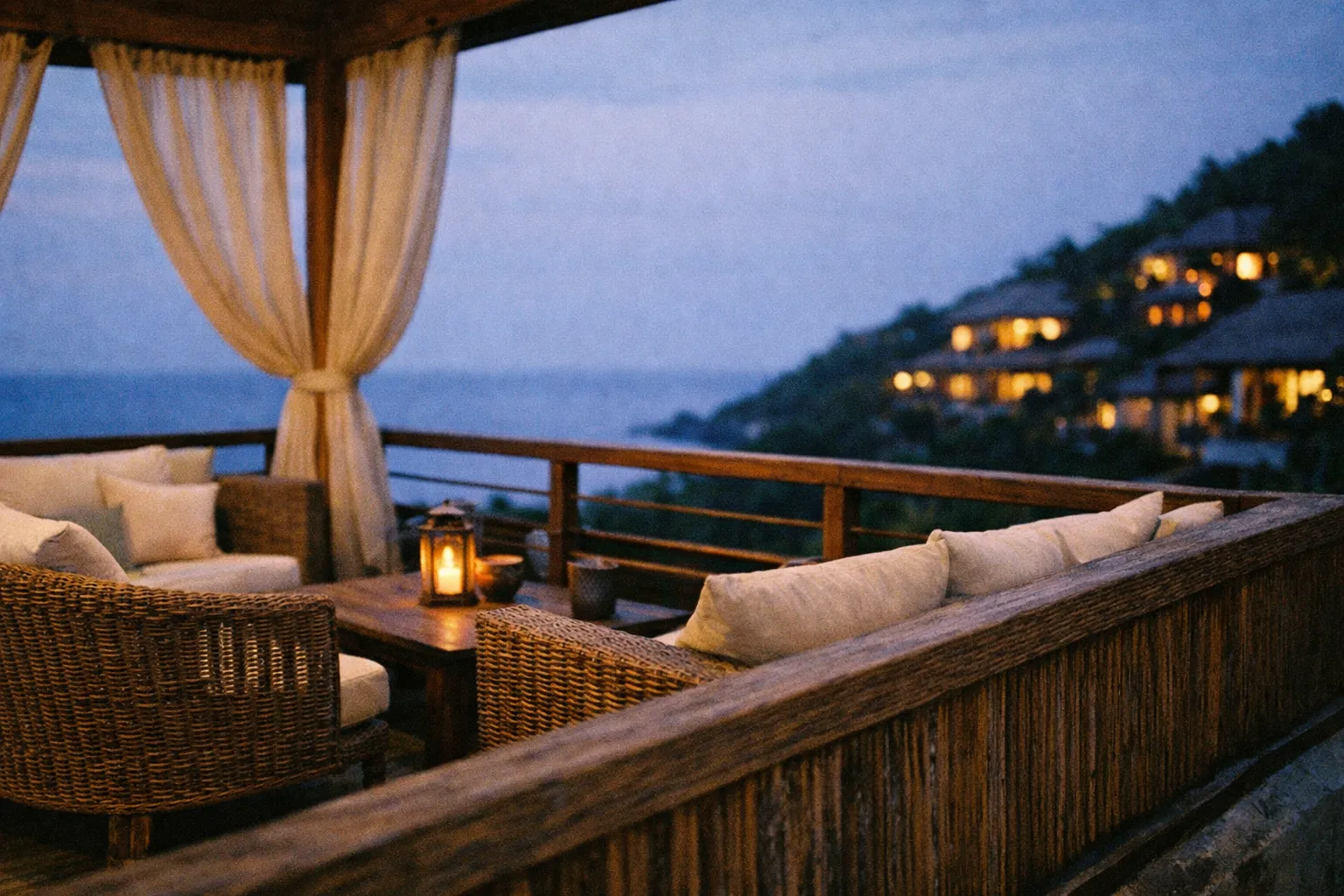 An outdoor lounge area with wicker furniture overlooks the ocean and hillside villas at dusk.