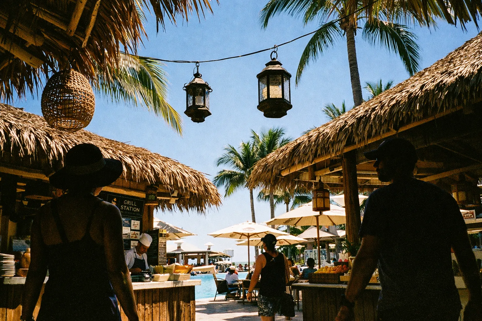 Guests walk between thatched-roof food stalls near a pool lined with palm trees and umbrellas.