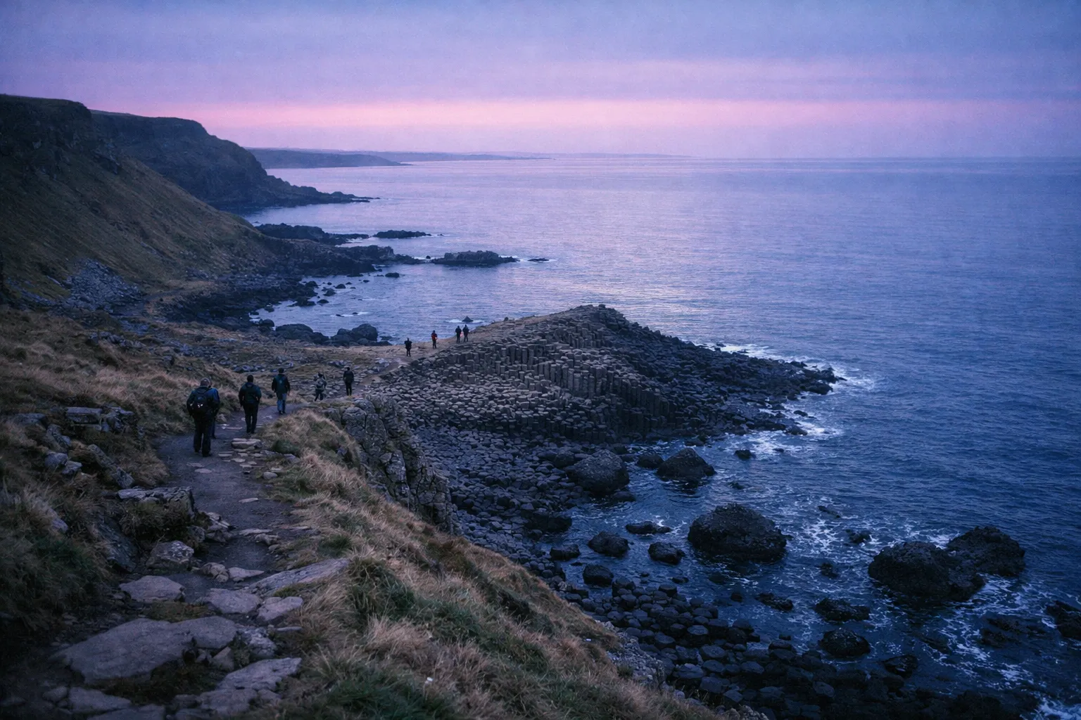 People walk along the coastal path near the basalt columns of Giant's Causeway at sunset.