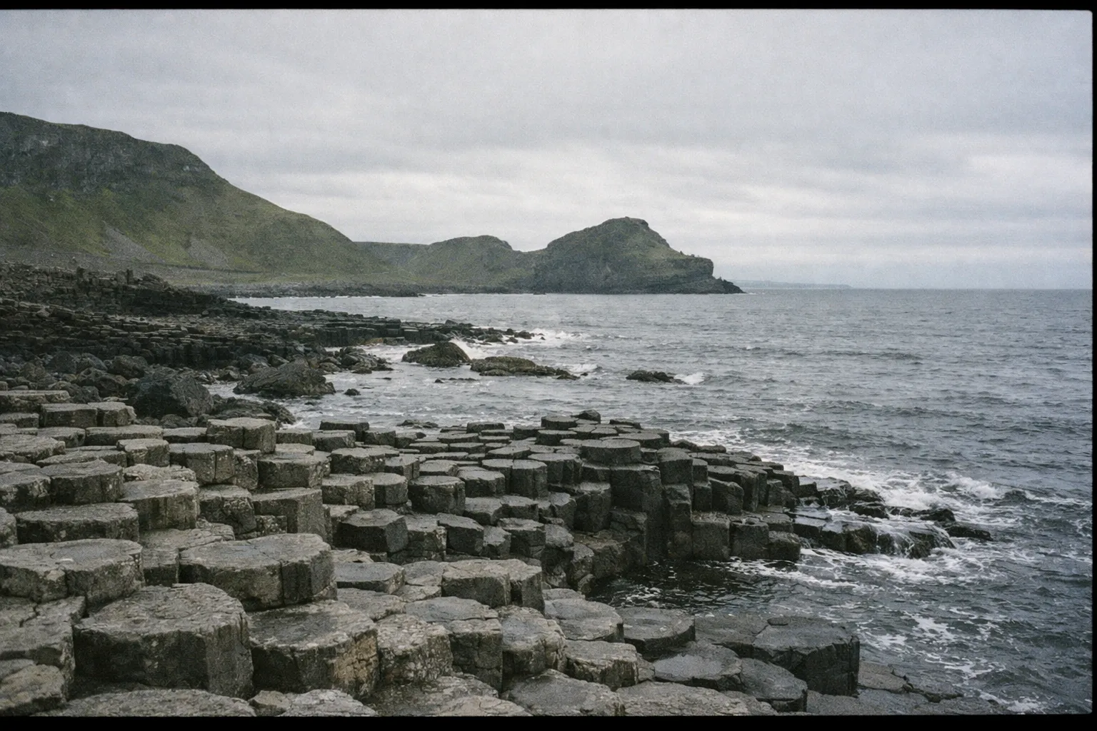 Basalt columns of the Giant's Causeway meet the sea under a cloudy sky with cliffs in the background.
