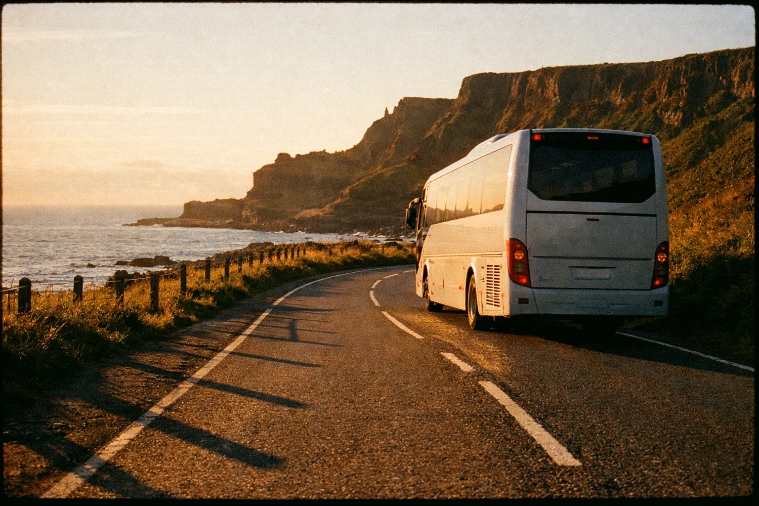 A tour bus travels along a coastal road near cliffs and the sea at sunset.