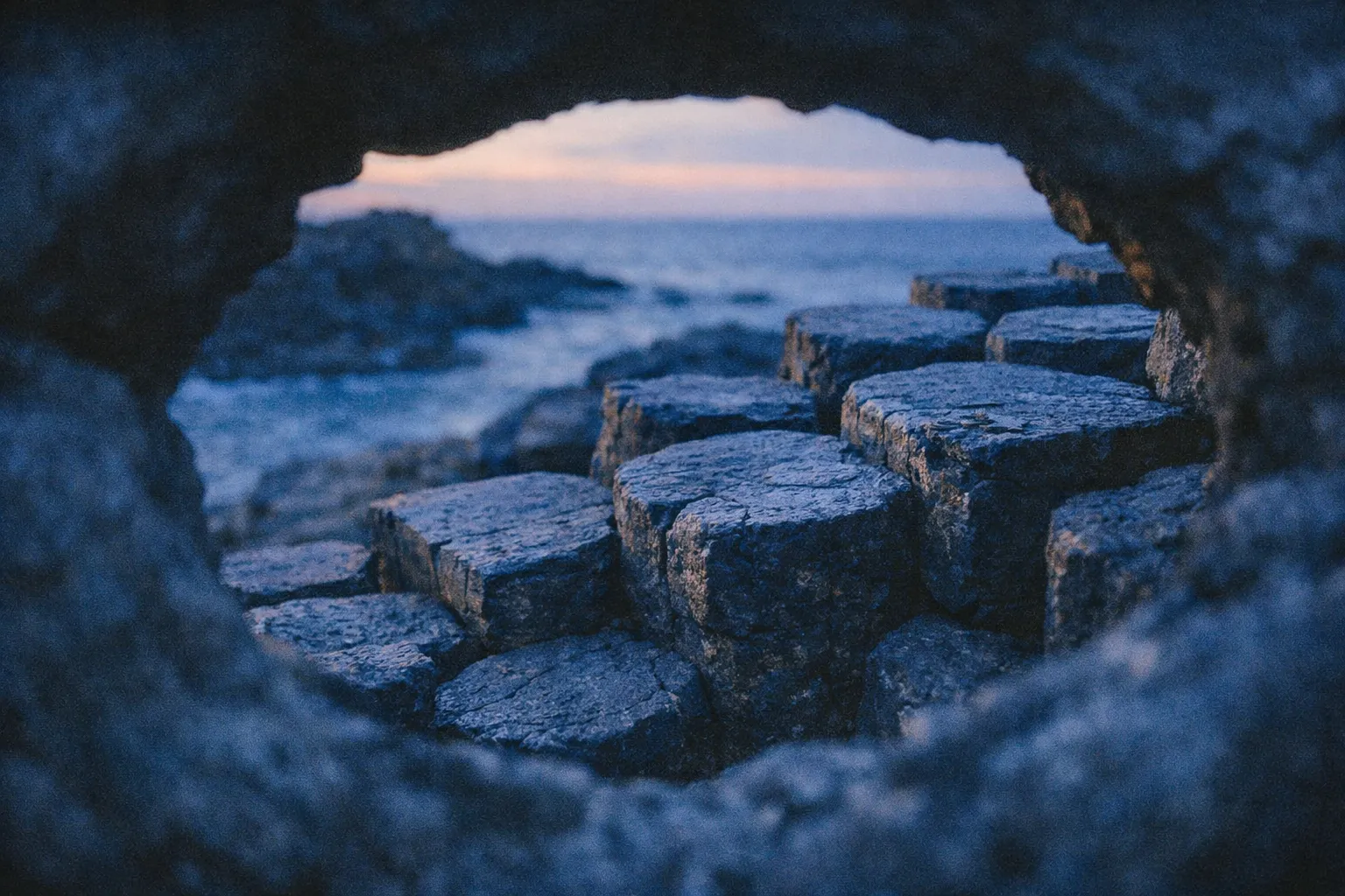 Hexagonal basalt columns at Giant's Causeway viewed through a rock opening at sunset by the sea.