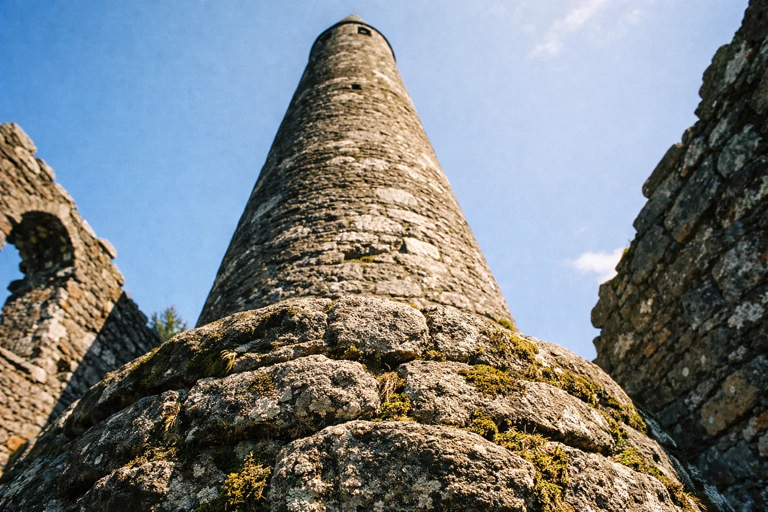 The round tower rises between old stone ruins under a clear blue sky.