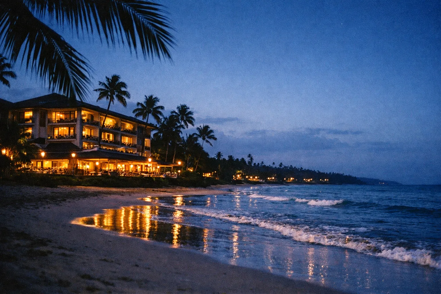 Beachfront hotel with palm trees at dusk, lights reflecting on the ocean and waves on the shore