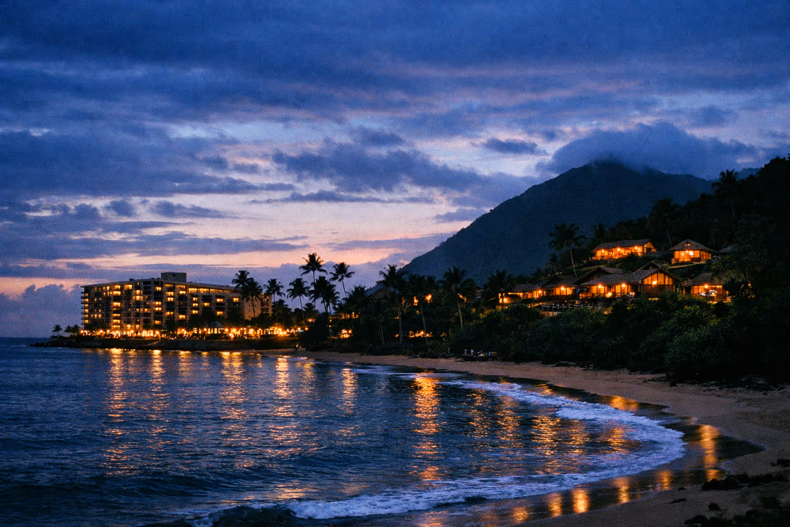 Beachfront resort buildings with lights on at dusk, palm trees and a mountain in the background