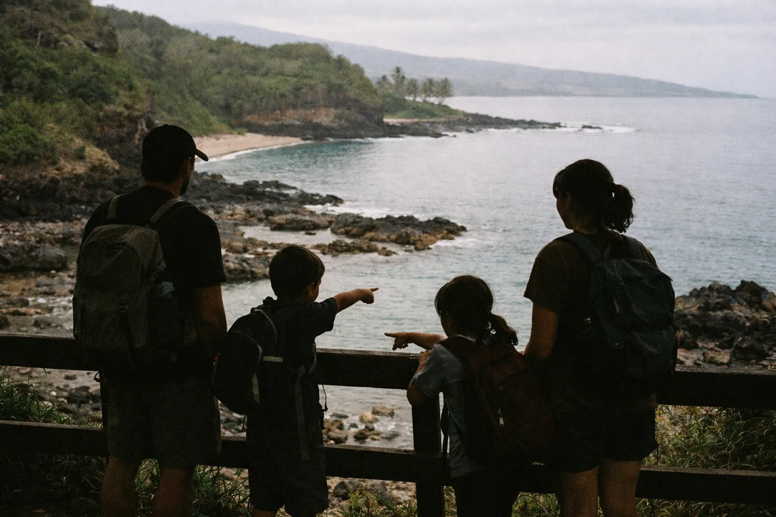 A family with backpacks looks out over a rocky beach and lush coastline in Hawaii.