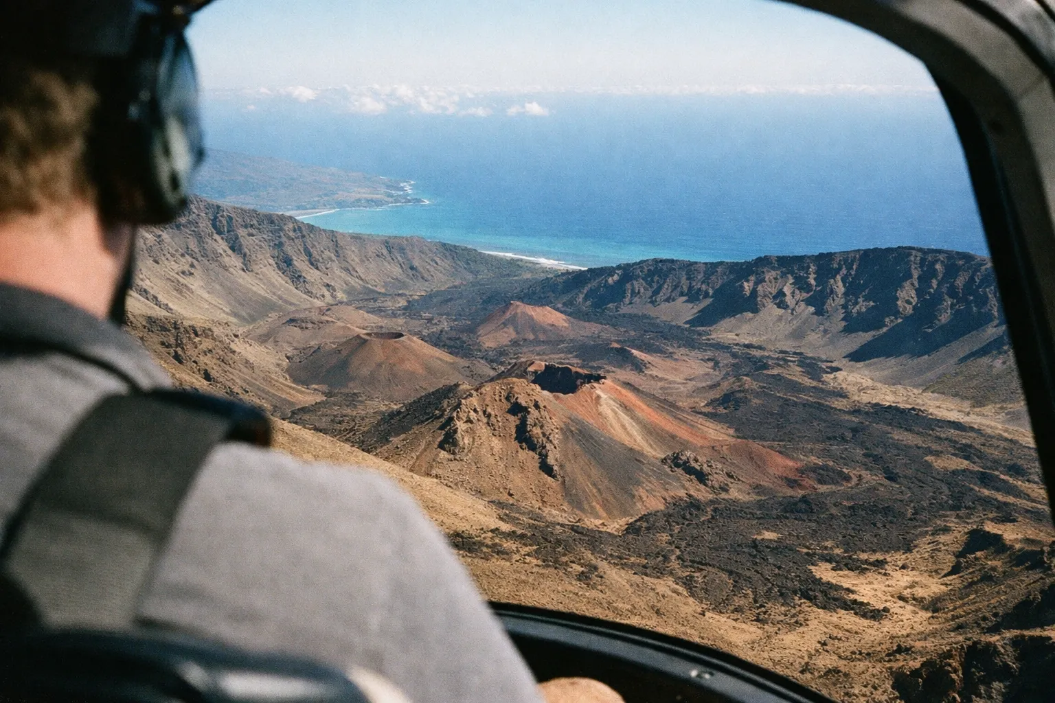 Person wearing headset looking out of helicopter window at volcanic landscape with coastline in the distance