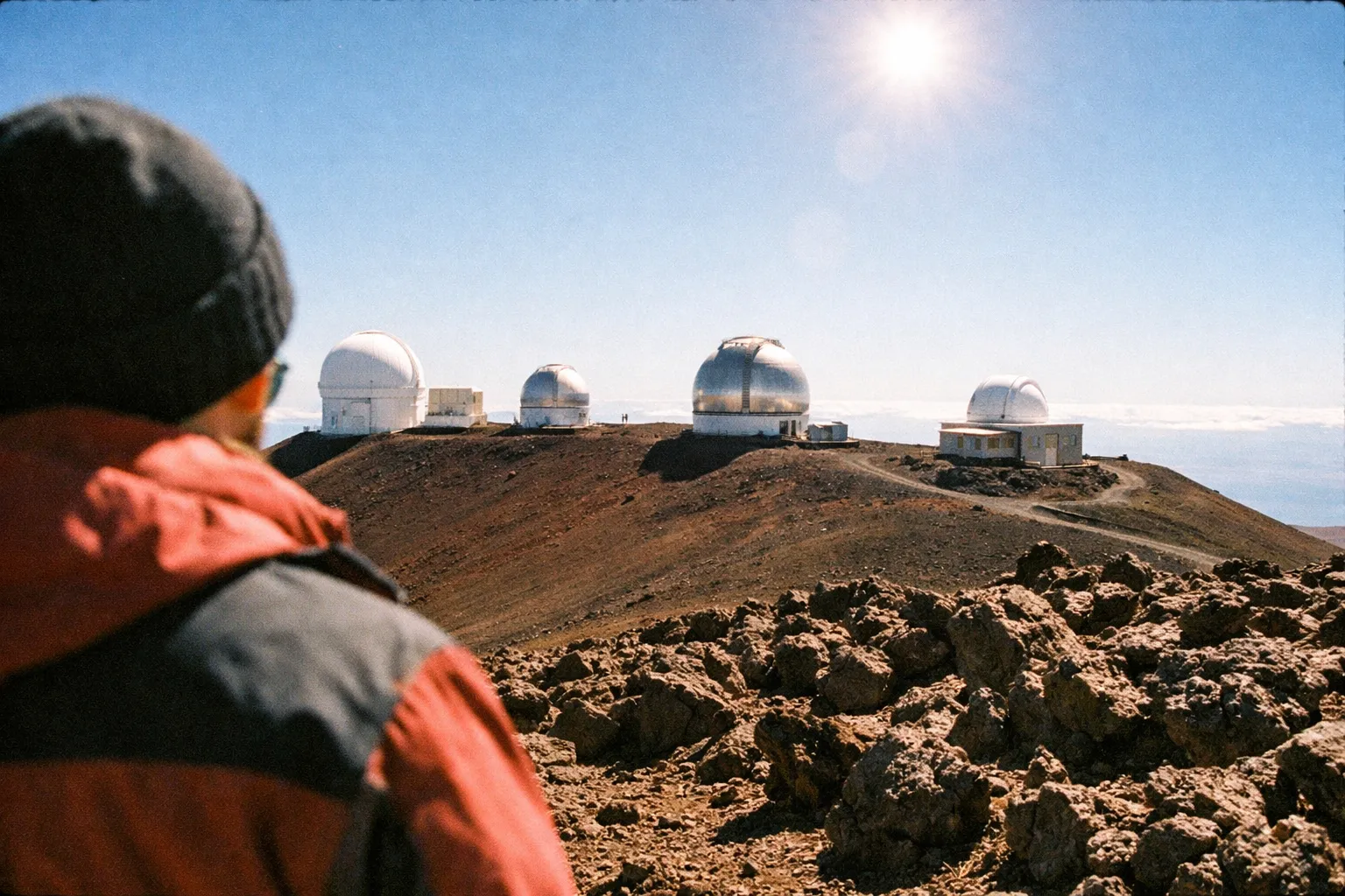 Person in warm clothing looking at observatory domes on a rocky hill under a bright sky