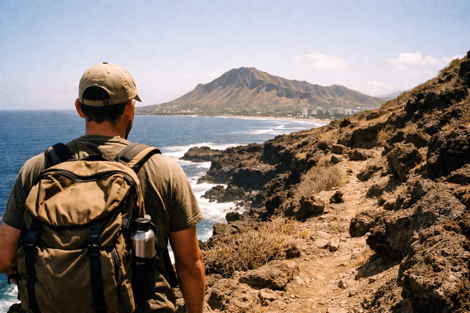 Person with a backpack standing on a rocky coastal trail overlooking the ocean and distant mountain