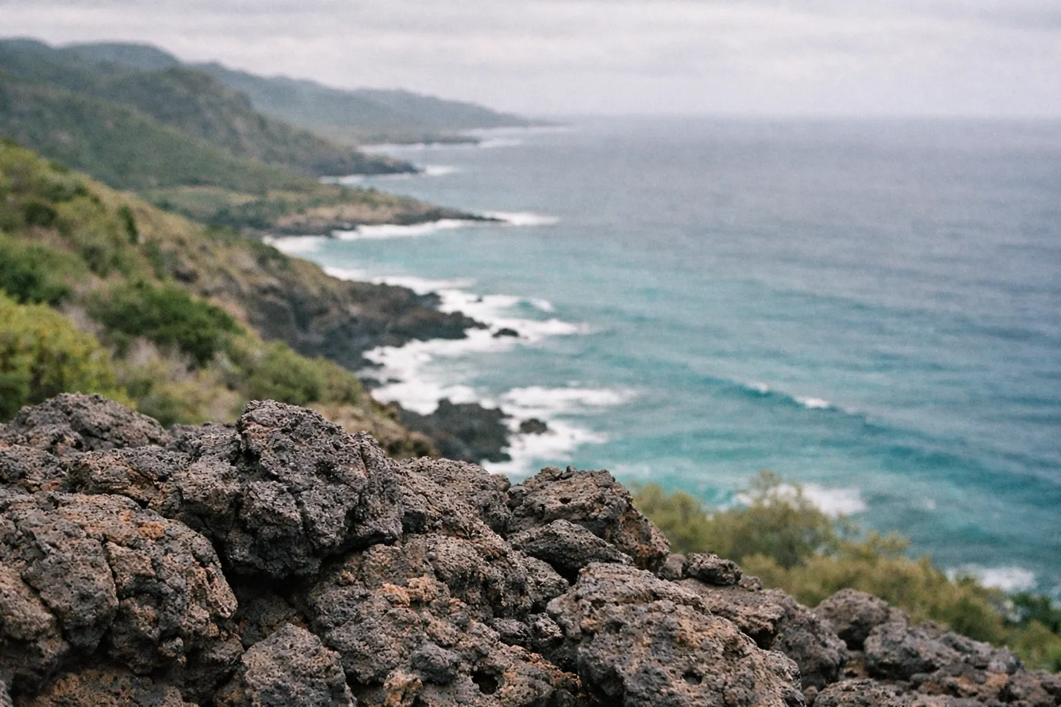 Rocky coastline with ocean waves and green hills under a cloudy sky