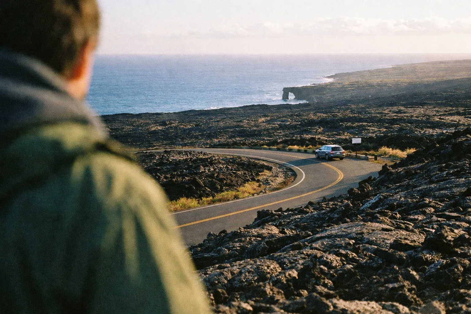 Over-the-shoulder view down a winding lava-field road toward coastal benches