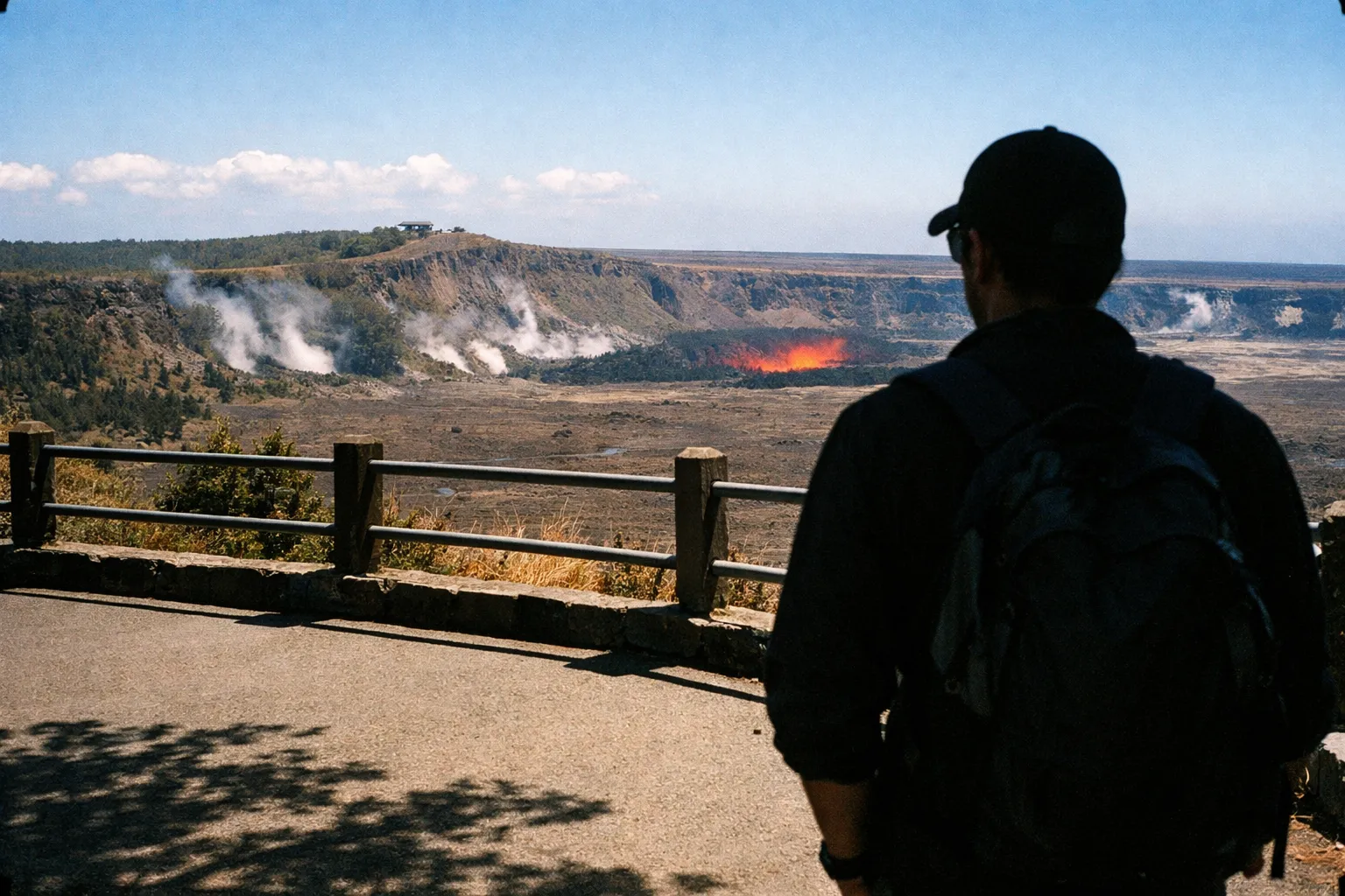 Over-the-shoulder view at a rim overlook with crater glow and distant plume