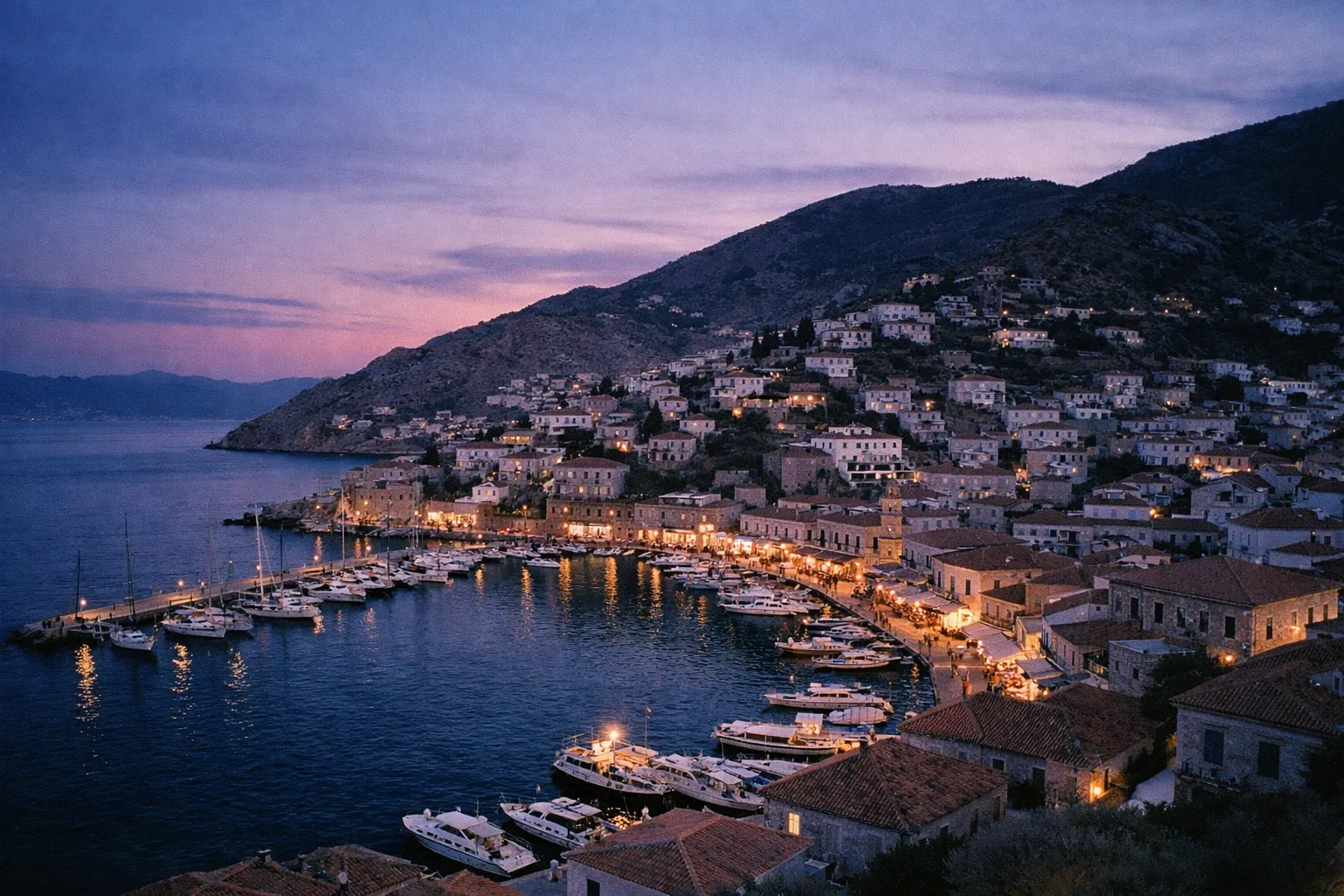 Hydra's harbor lined with boats and stone buildings at dusk, nestled against a hillside in Greece.