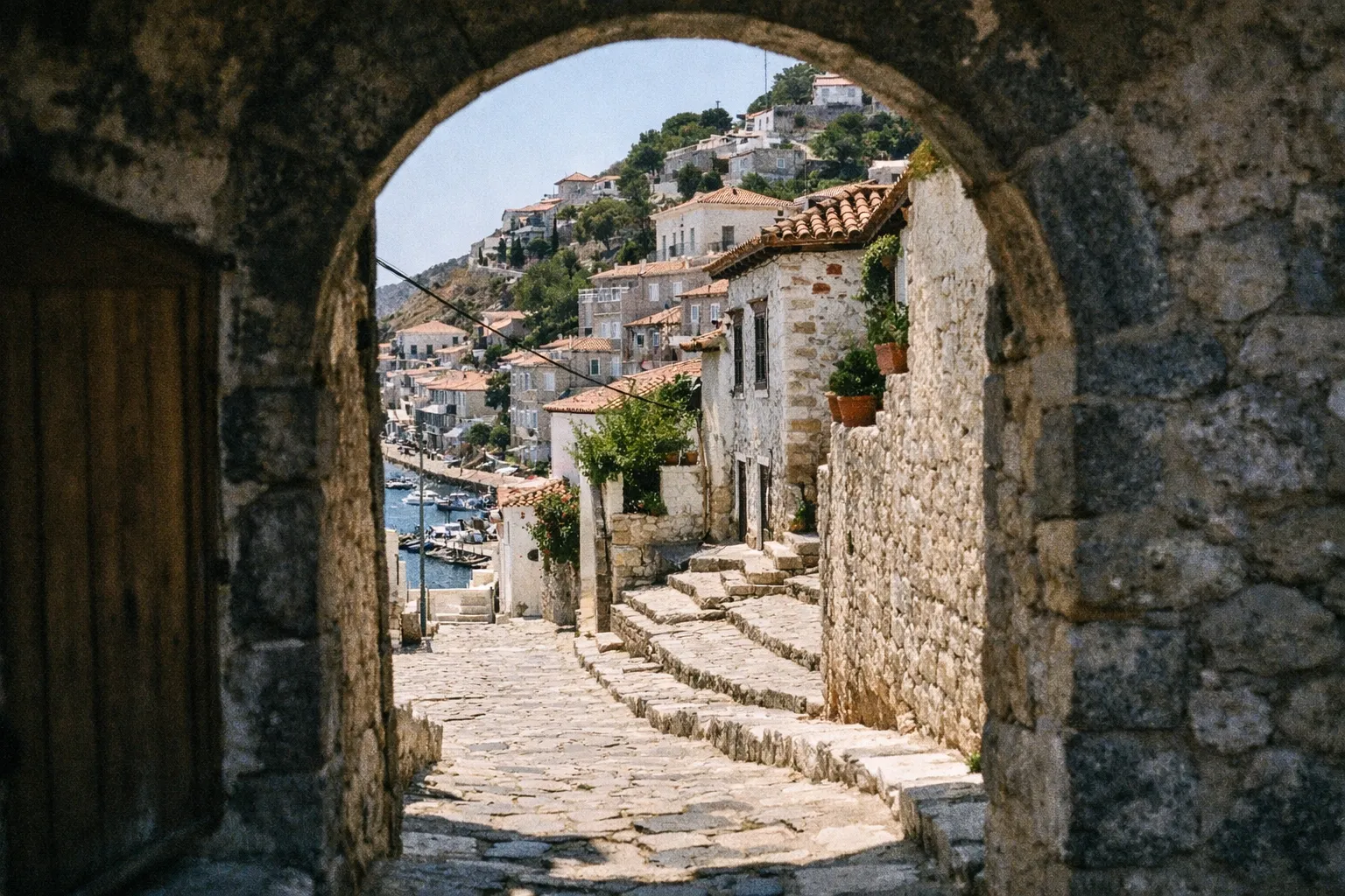 A stone archway frames a narrow cobblestone street leading down to the harbor in Hydra, Greece.