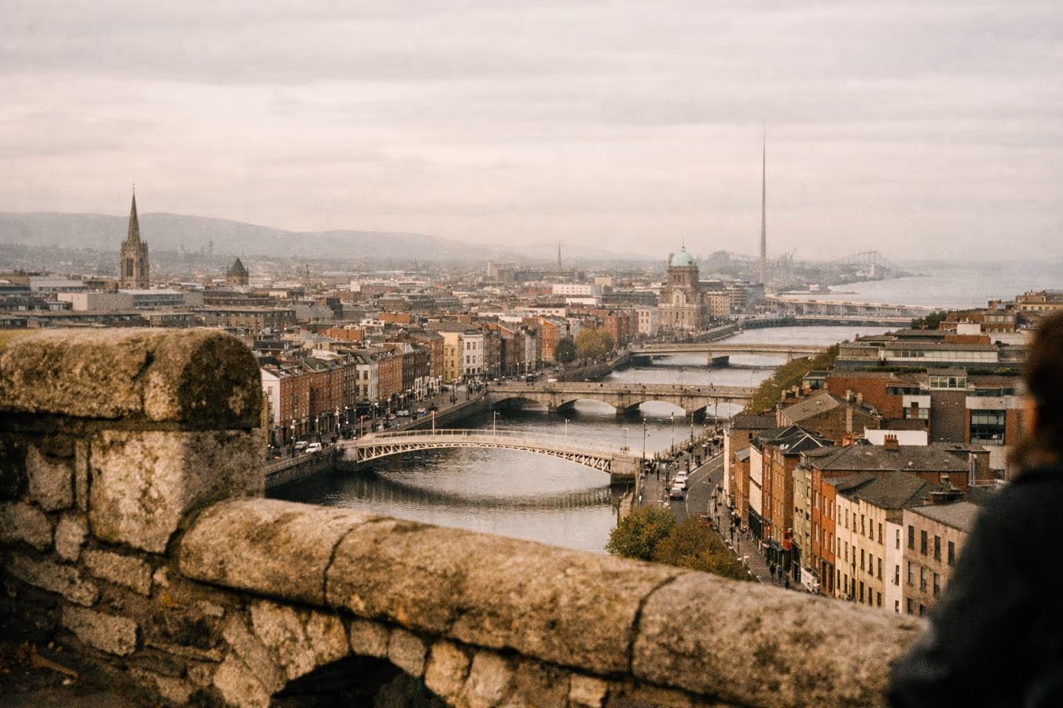 View over central Dublin with the River Liffey, bridges, and city landmarks visible under a cloudy sky.