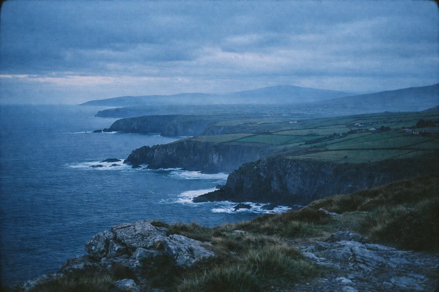 Rocky cliffs and patchwork fields along Ireland's Atlantic coastline beneath a cloudy sky at dusk.