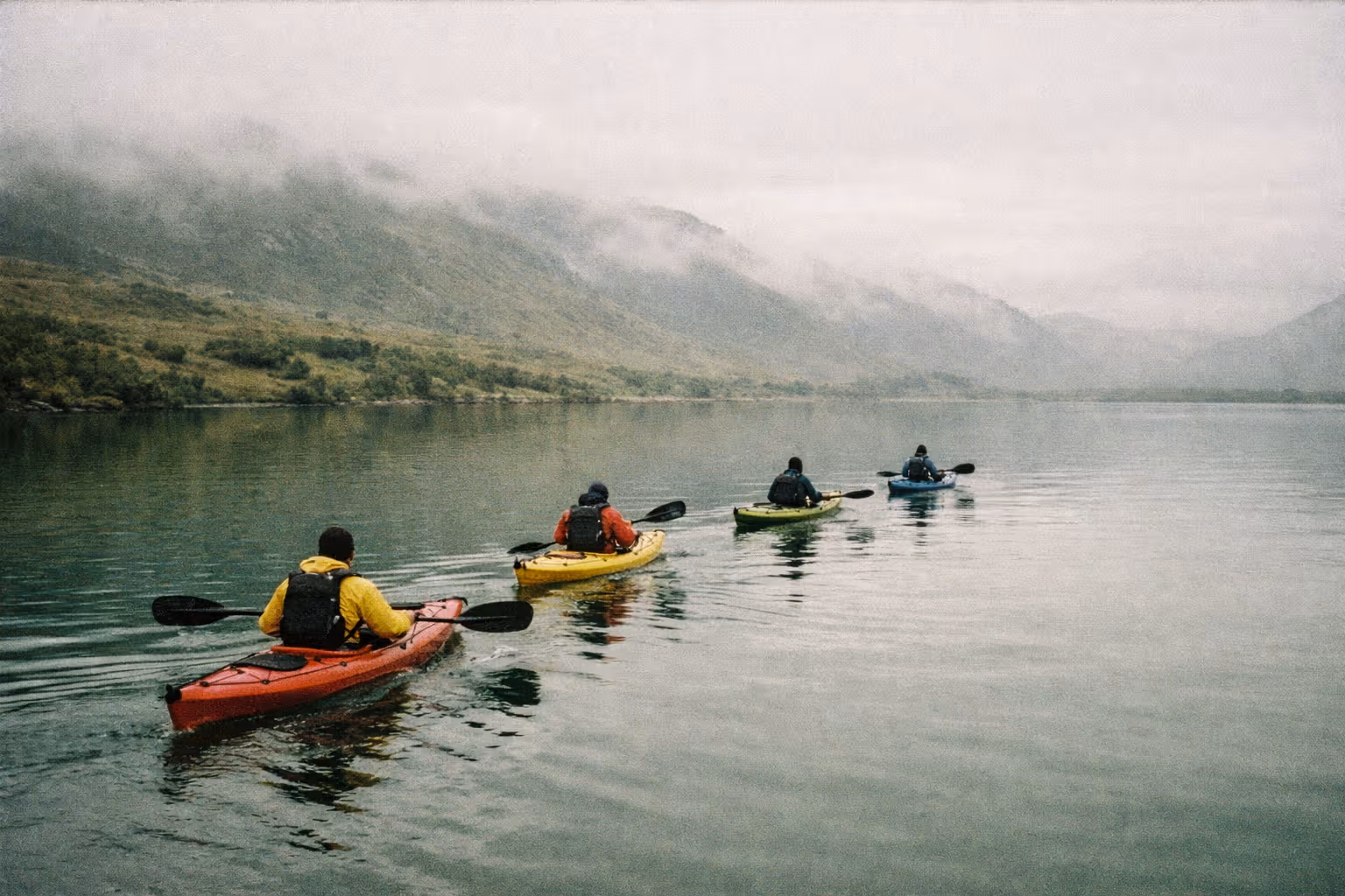 Four people kayaking on a calm lake surrounded by misty green hills.