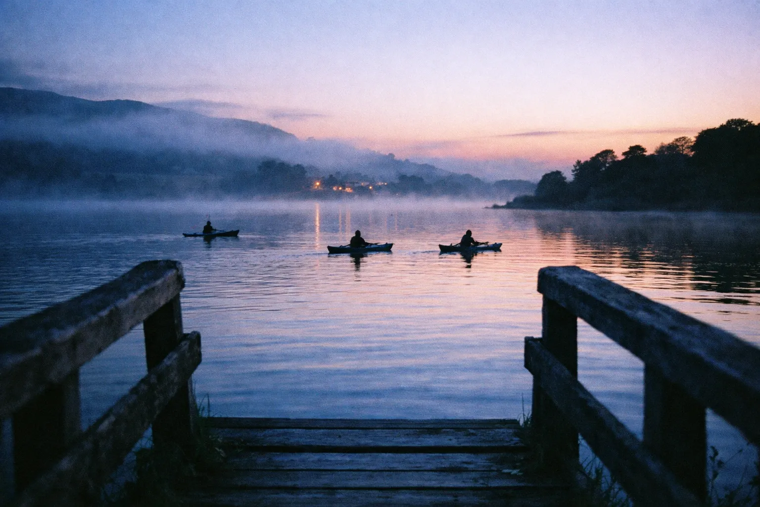 Three people kayak on a calm lake at dusk, viewed from a wooden pier with misty hills in the background.