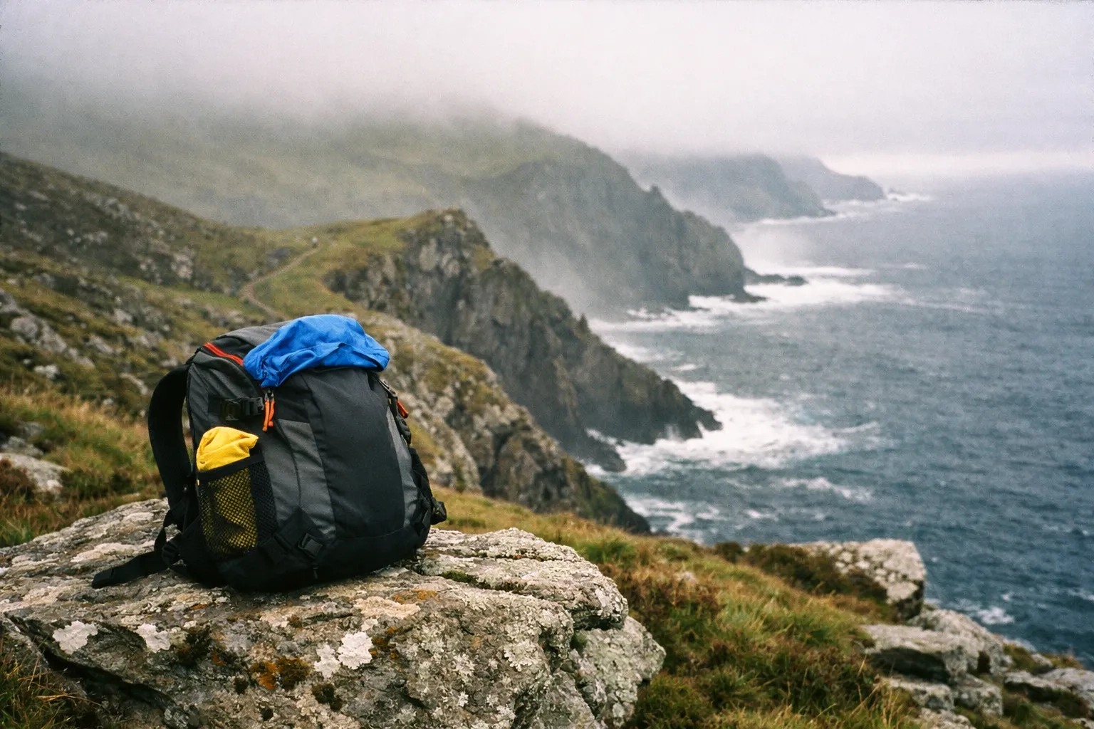 A backpack sits on a rocky cliff overlooking the misty coastline of Ireland.