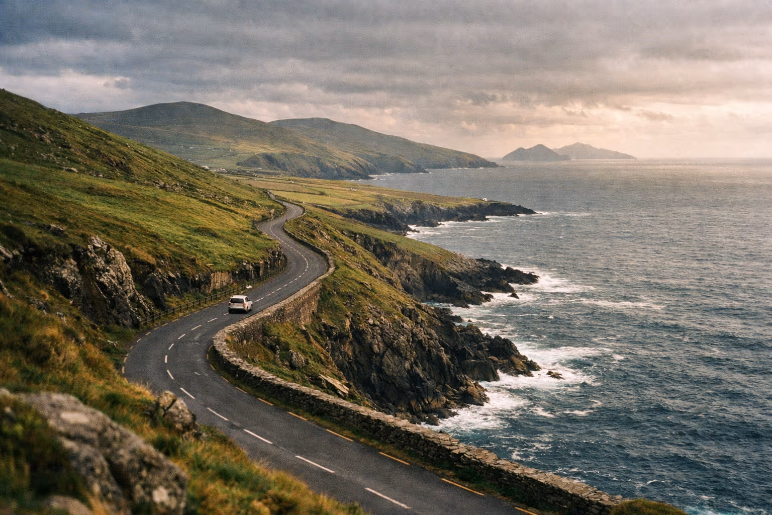 A car drives along a winding coastal road on the Dingle Peninsula in Ireland, with cliffs and ocean below.