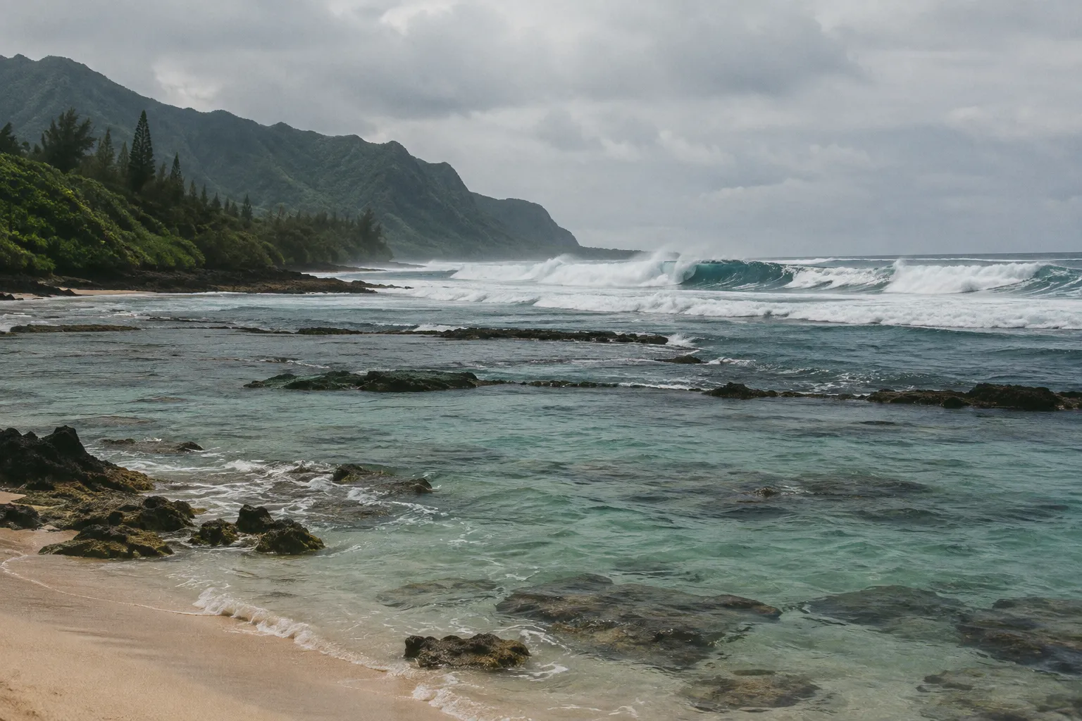 Rough surf breaks over a rocky reef beside a beach beneath Kauai's green coastal mountains.