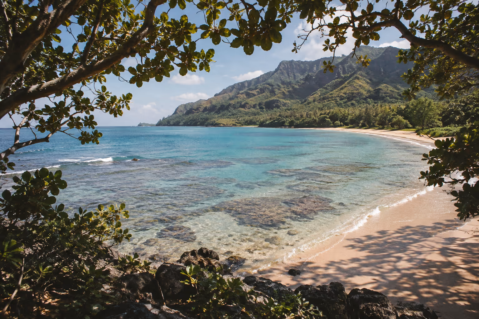 Clear shallow reef water meets a sandy north shore beach on Kauai, framed by trees and green mountains.
