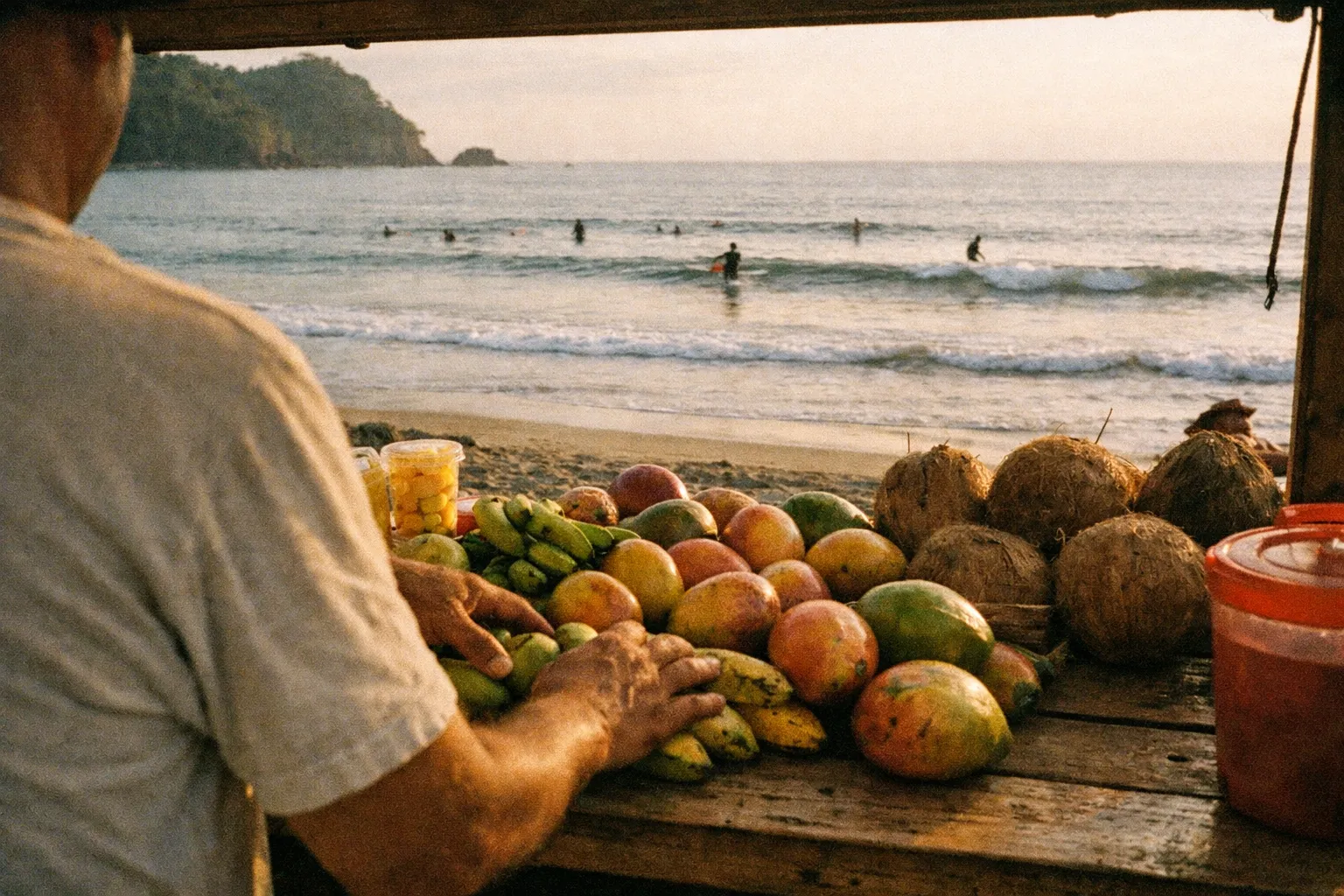 A vendor arranges fresh fruit at a beachside stall near the ocean with swimmers in the background.