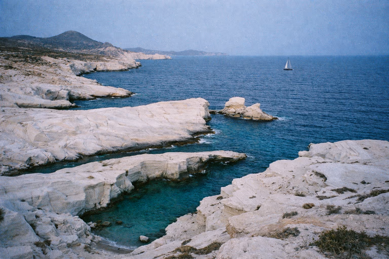 White rock formations and turquoise waters of Sarakiniko Beach on Milos with a sailboat in the distance.