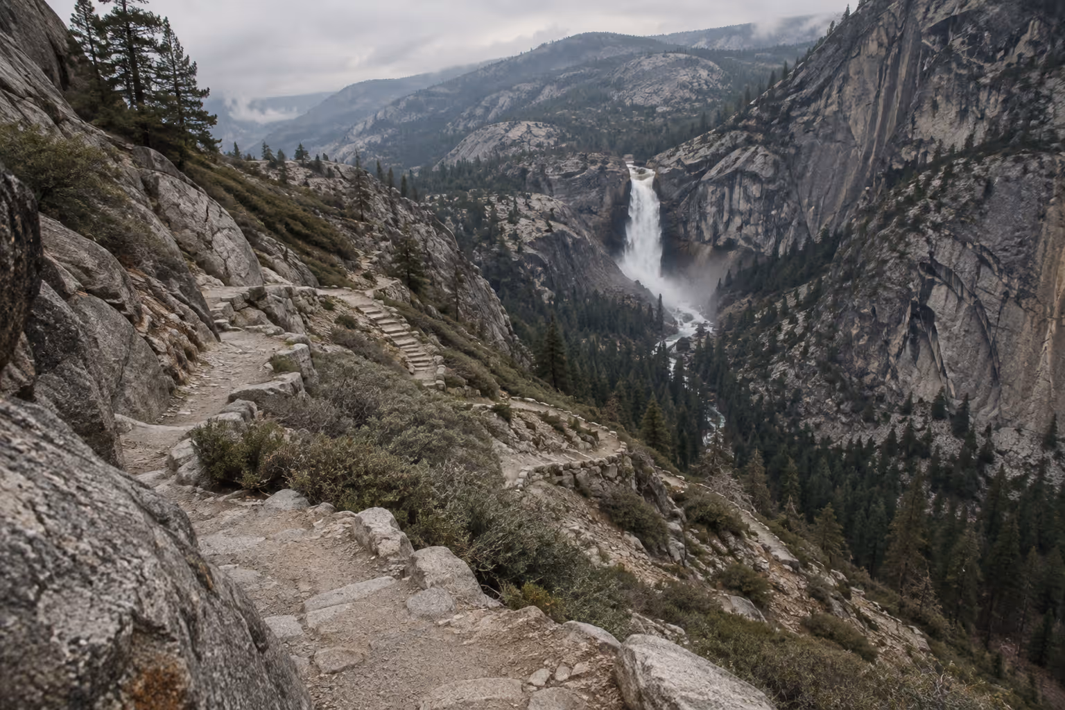 Rocky trail with stone steps winding along a granite slope above a forested canyon, a white waterfall in the distance