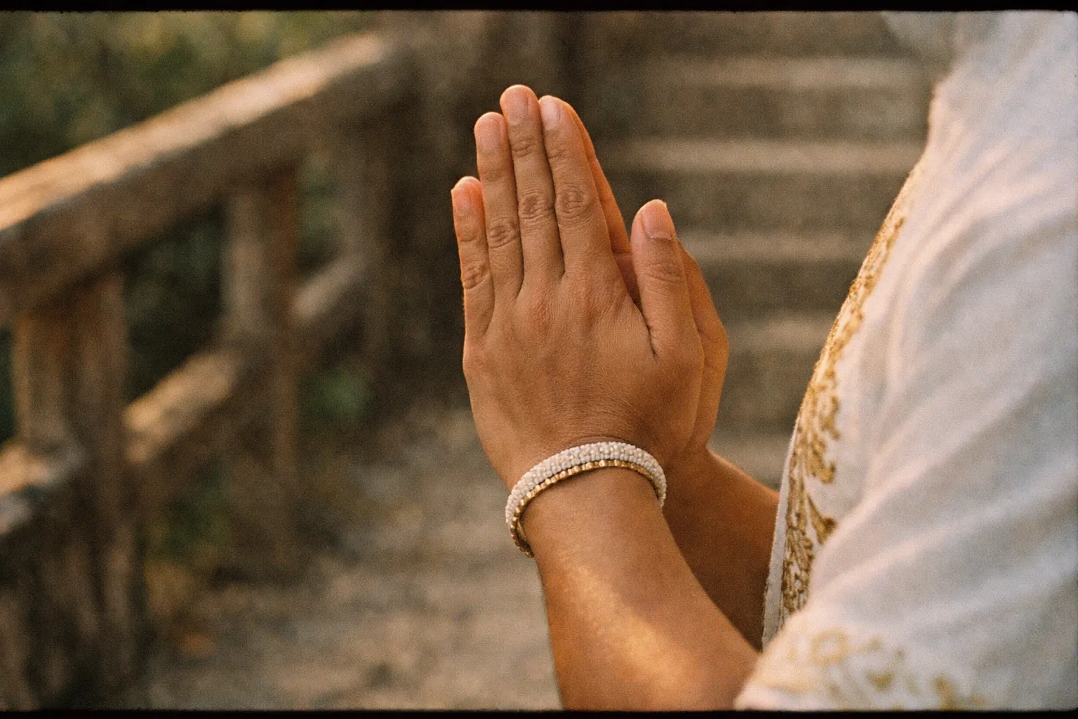 Hands in a Balinese greeting (palms together) seen from the side against a wooden railing at sunset, no faces visible