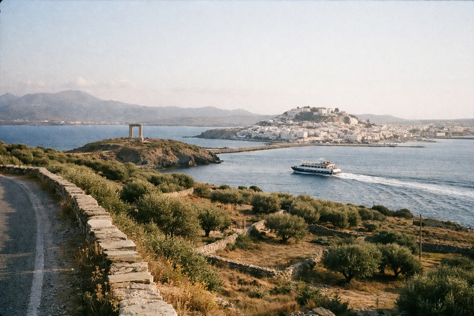 The Portara ruins on a small islet near Naxos town, with a ferry in the sea and olive groves in the foreground.