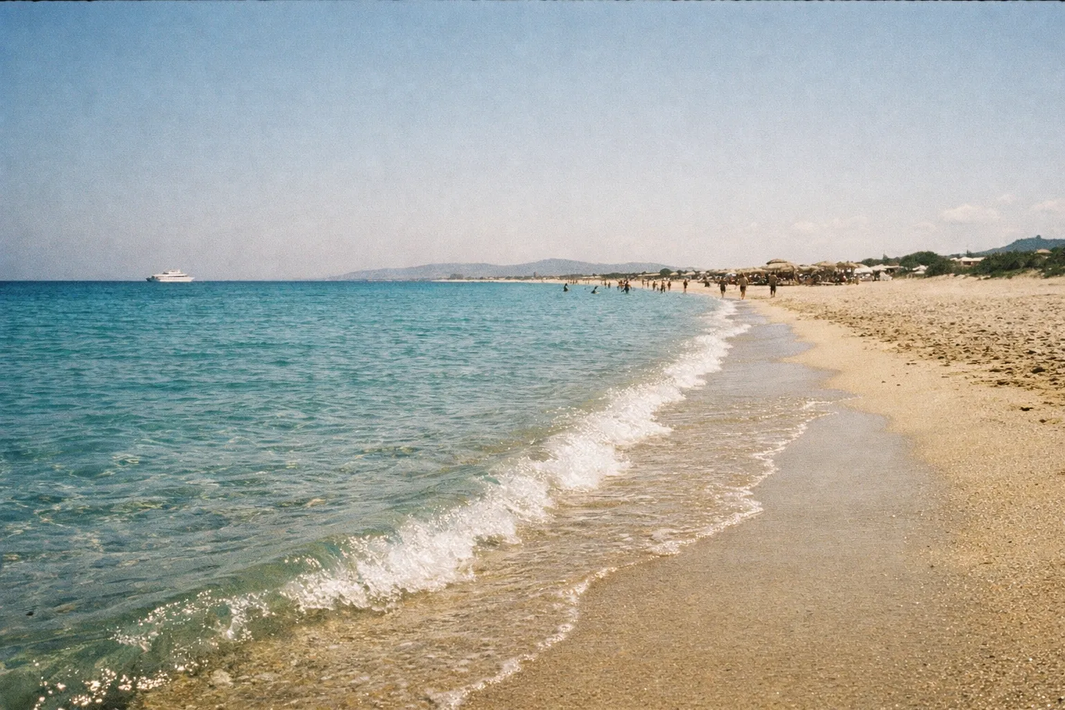 People walk and relax along a sandy beach with clear blue water and a distant ferry near Naxos, Greece.