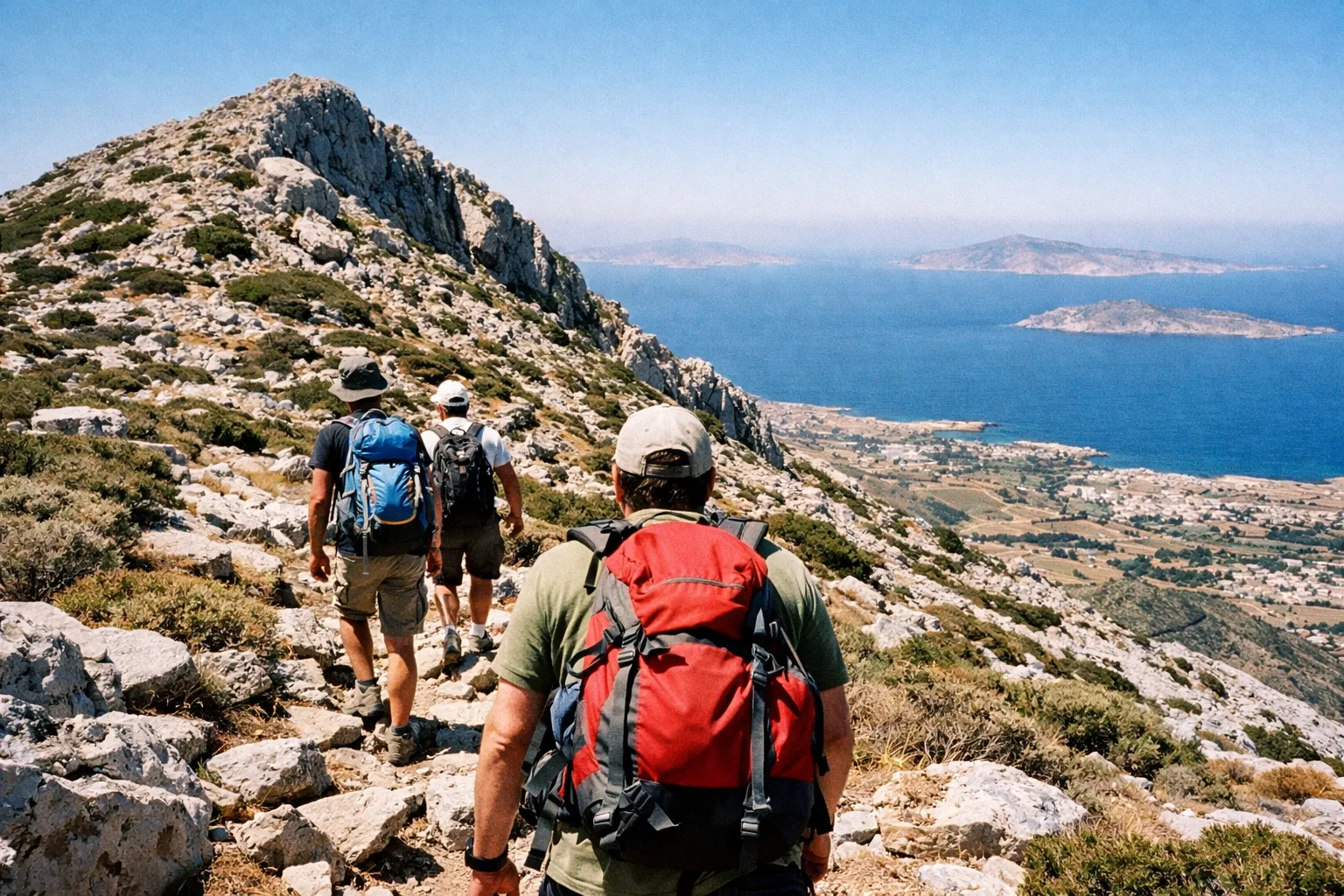 Hikers with backpacks walk up a rocky mountain trail overlooking the sea and villages on Naxos, Greece.