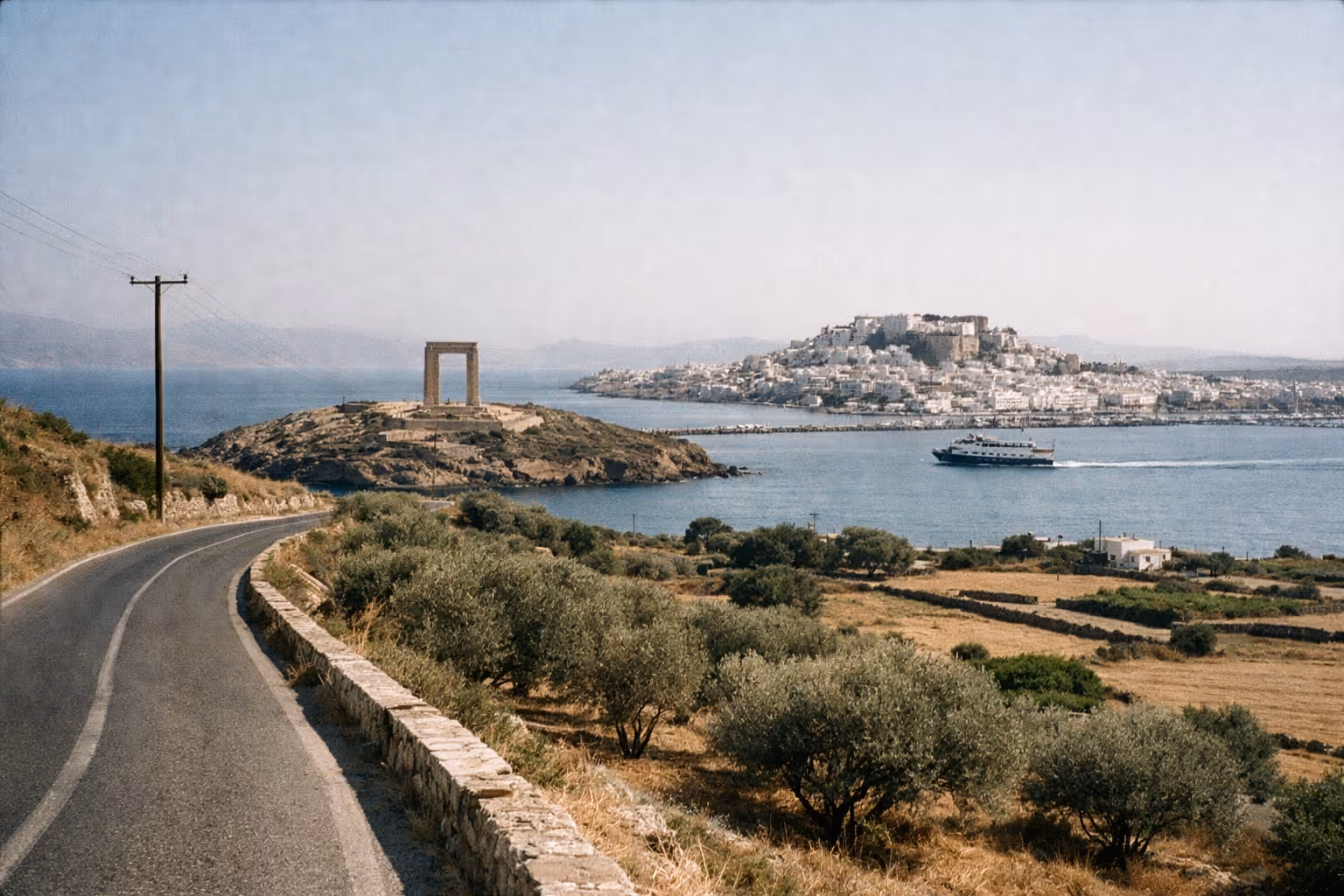 The Portara stone gateway sits on a small islet near the port town of Naxos with a ferry in the bay.