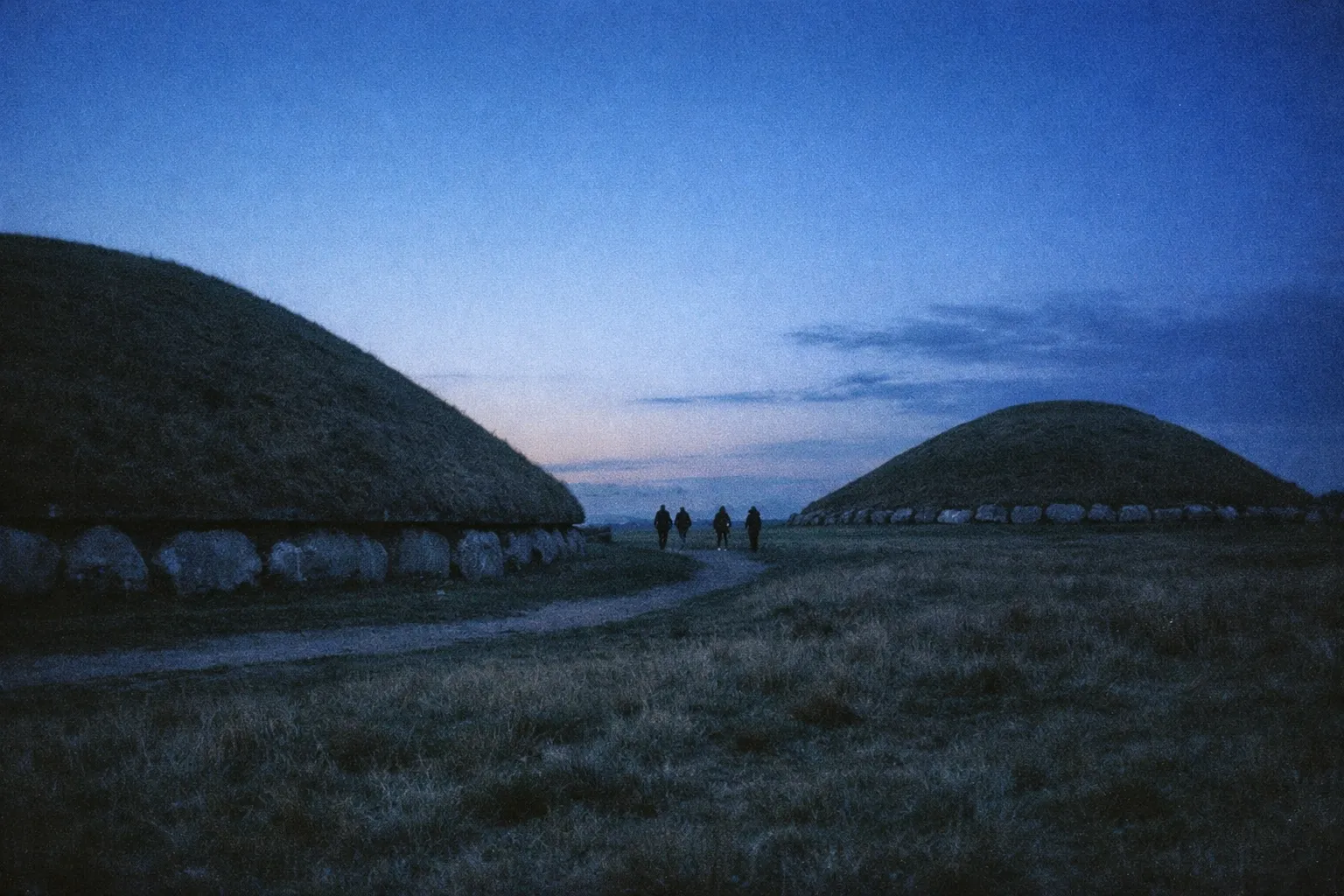 People walk along a path between two large ancient mounds at dusk in the Boyne Valley.