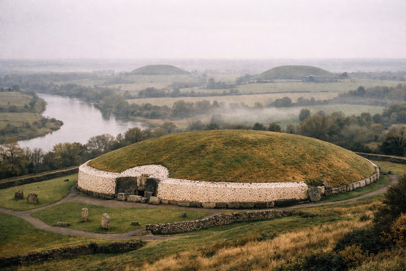 The Newgrange passage tomb sits on a hill overlooking the Boyne Valley and river in a misty rural landscape.