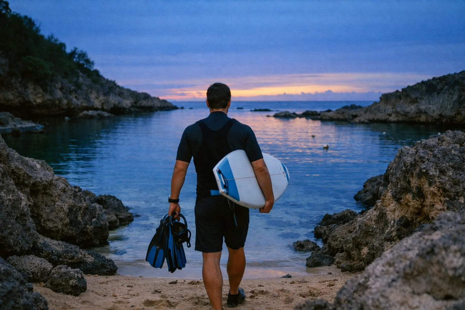 A man holding a surfboard and fins walks toward a rocky beach cove at sunset.