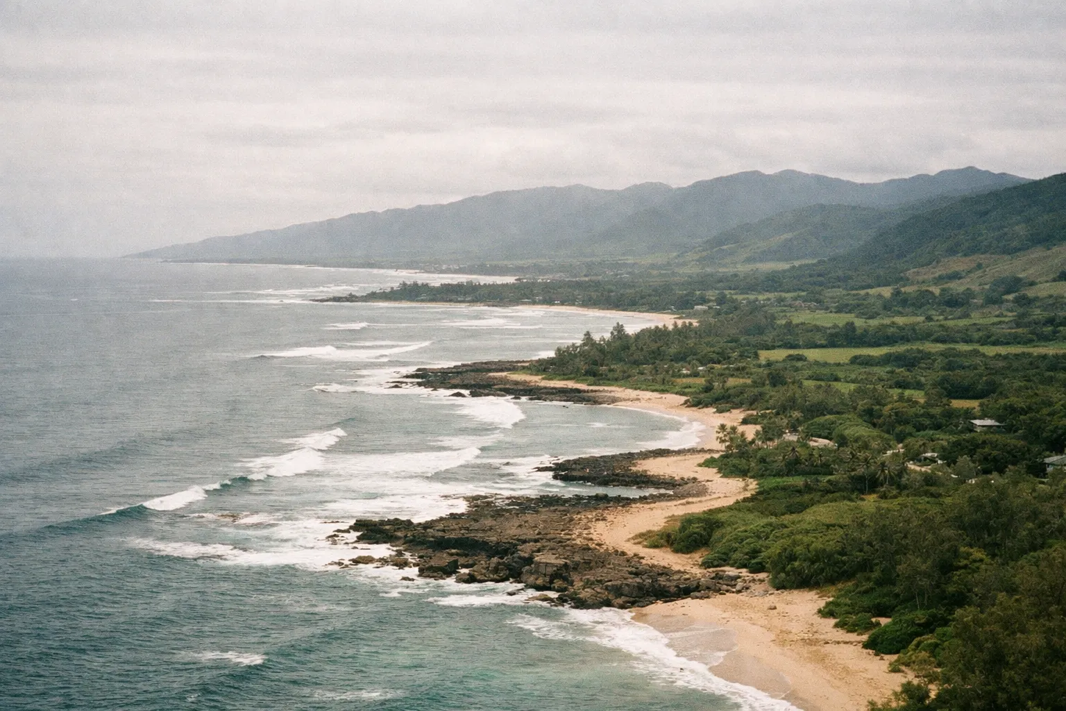 Waves break along a sandy, rocky shoreline bordered by lush green mountains on the North Shore of Oahu.