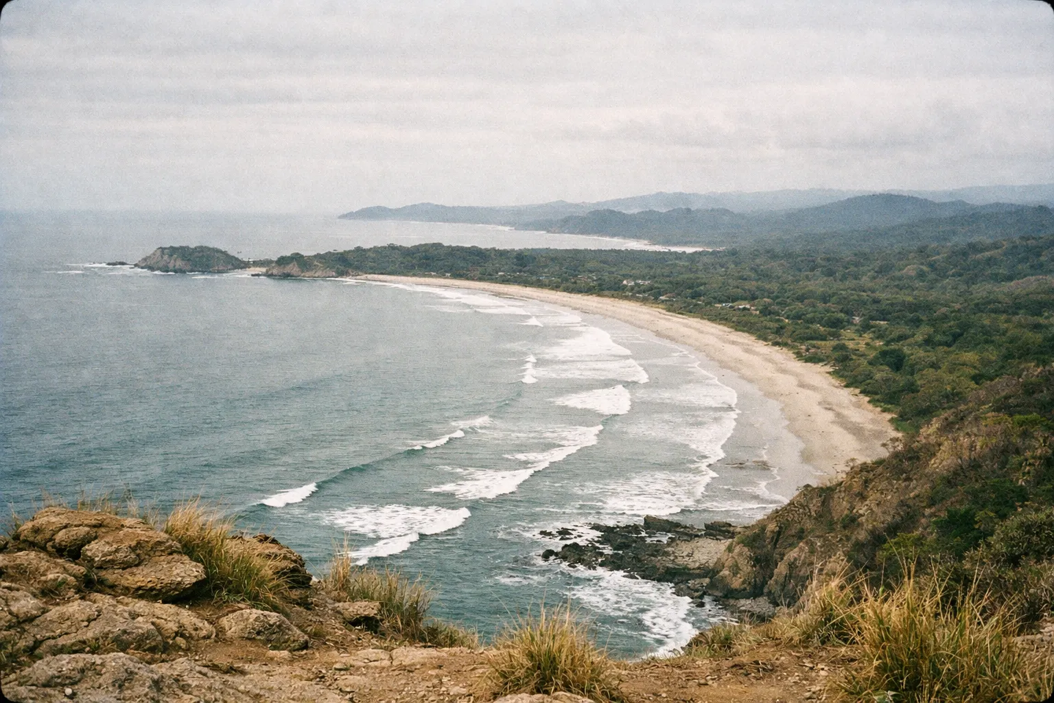 A wide sandy beach curves along the coast with waves and forested hills in Nosara, Costa Rica.