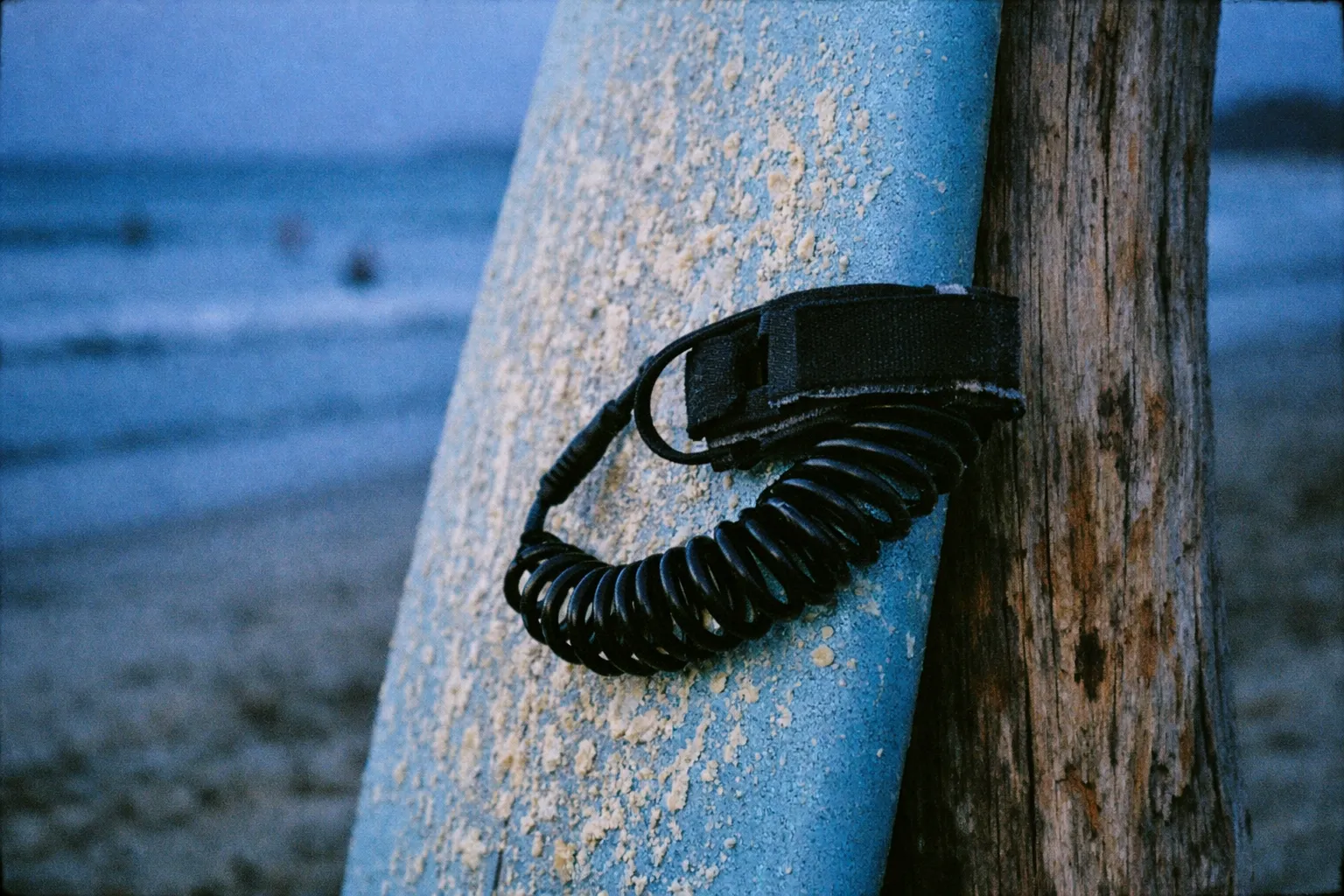A close-up of a sandy surfboard with a leash leaning against a wooden post on a beach.