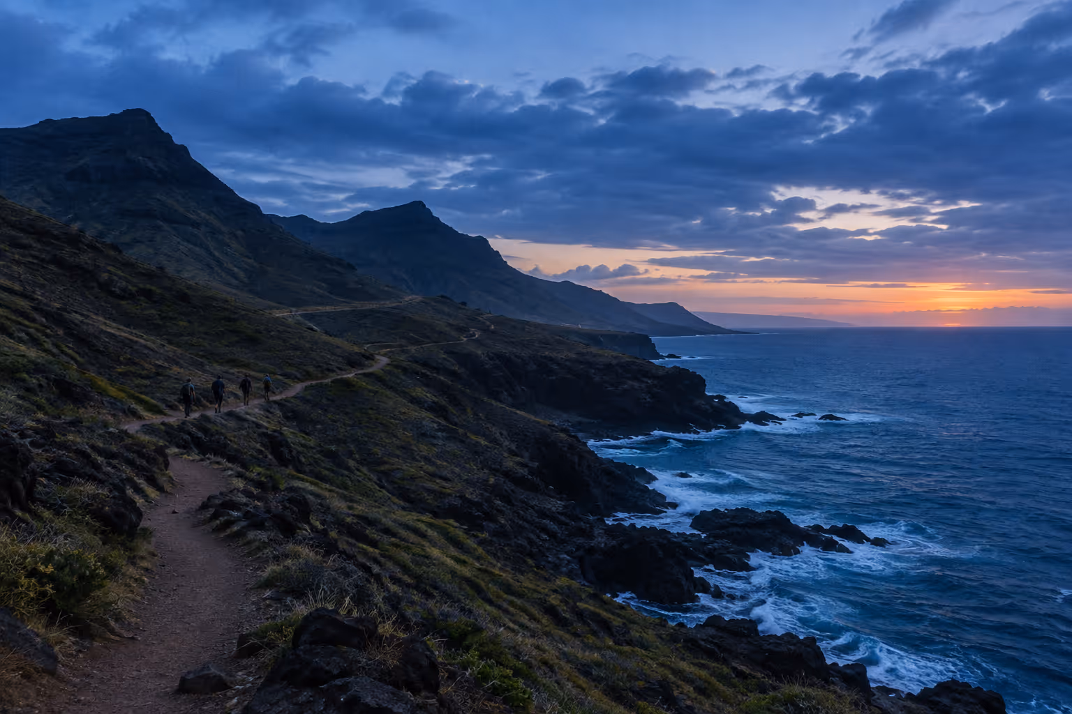 Hikers follow a rocky coastal trail above the ocean at sunset.