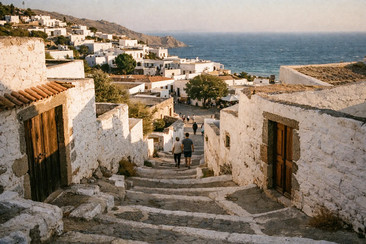 People walk down stone steps between whitewashed houses in Chora, Patmos, with the sea in the background.
